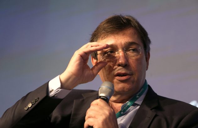 Um homem de óculos e terno falando ao microfone, com a mão na altura dos olhos. Usa um cordão Credit Suisse.