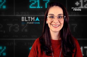 Mulher sorridente com óculos em frente a uma tela digital exibindo dados financeiros e logos "investnews".