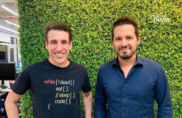 Dois homens sorrindo, um com camiseta de programação, à frente de parede de folhagem verde.