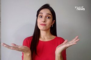 Mulher de camisa vermelha encolhe os ombros com expressão de incerteza e mãos abertas.