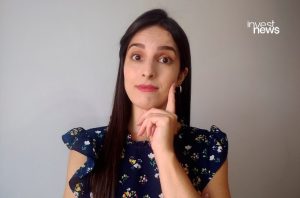 Mulher de cabelo escuro e blusa floral azul, dedo na bochecha com expressão pensativa. Fundo cinza liso.