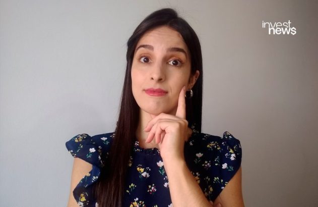 Mulher de cabelo escuro e blusa floral azul, dedo na bochecha com expressão pensativa. Fundo cinza liso.