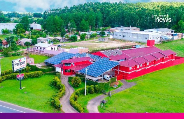 Aéreo: Motel Pepper vermelho com telhados cheios de painéis solares, cercado por paisagem verde.