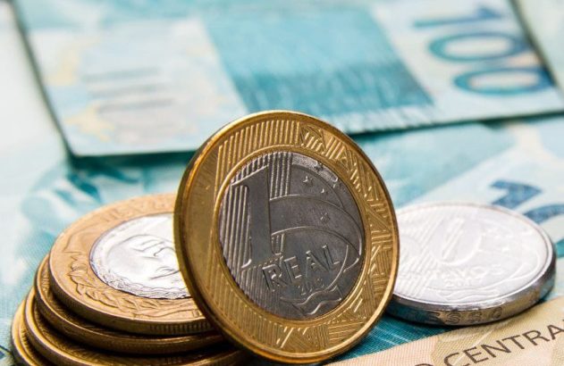 Moedas de Real e cédulas brasileiras. Uma moeda de 1 Real em pé sobre notas de 50 e 100 Reais.