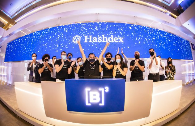 Grupo de pessoas com máscaras celebra lançamento da Hashdex na B3. Telão azul com logo e pontos brancos ao fundo.