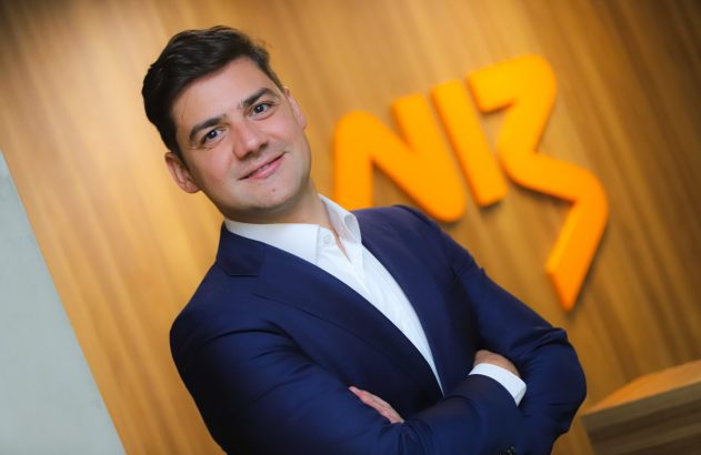 Homem sorridente de terno azul com braços cruzados, em frente a uma parede de madeira com logo laranja.