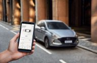 Mão segura smartphone com app de carro exibindo status e autonomia, com carro cinza desfocado ao fundo.