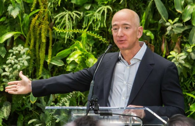 Jeff Bezos discursa e gesticula em pódio. Ao fundo, parede de plantas.