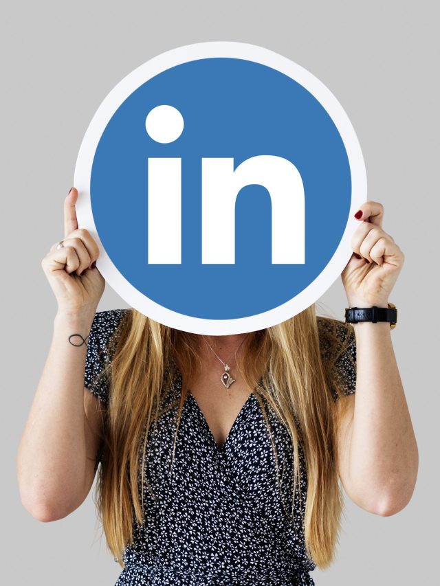 Pessoa com cabelos loiros segurando um cartaz azul do LinkedIn que cobre seu rosto, em frente a um fundo cinza.