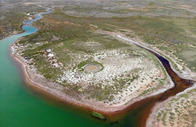 Vista aérea de encontro de rios em paisagem árida. Um rio claro e outro escuro desaguam em lago verde. Vegetação esparsa.