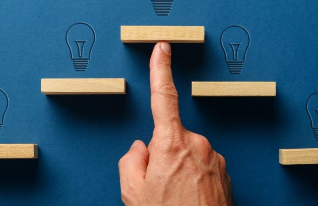 Mão impulsiona bloco de madeira para acender lâmpada em fundo azul. Outras lâmpadas e blocos sugerem uma escada de ideias.