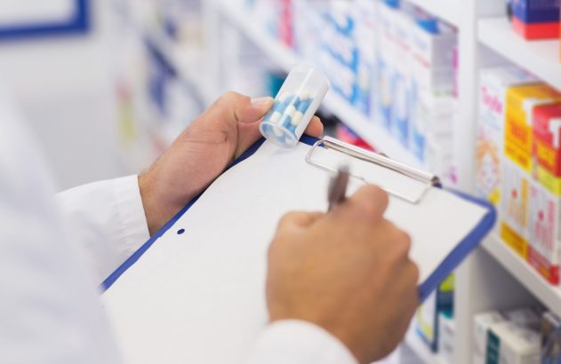 Pessoa segura recipiente com pílulas e escreve em prancheta, com prateleiras de remédios ao fundo.