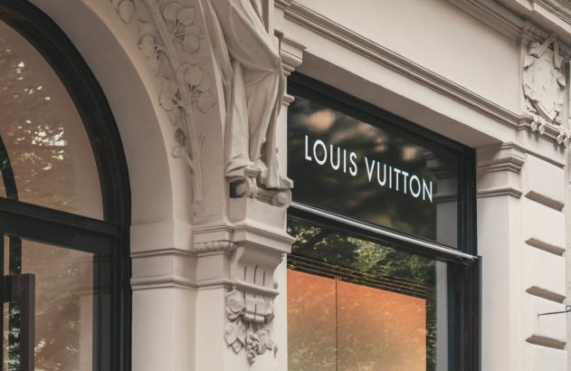 Detalhes da fachada Louis Vuitton: ornamentos, estátua e janelas amplas com reflexos de árvores no vidro.