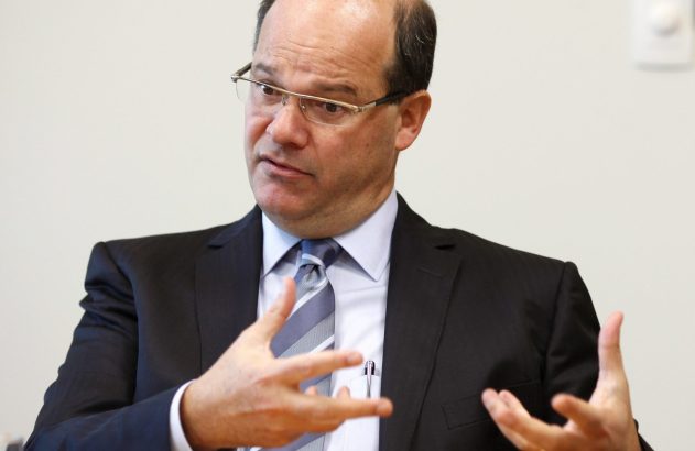 Homem de óculos, terno e gravata, gesticula com as mãos abertas enquanto fala. Fundo claro.