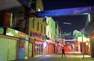 Rua noturna com lojas vibrantes, letreiros neon de 'Beach Club', 'DOLLHOUSE' e 'Döner-Freiheit'. Pessoa caminha.