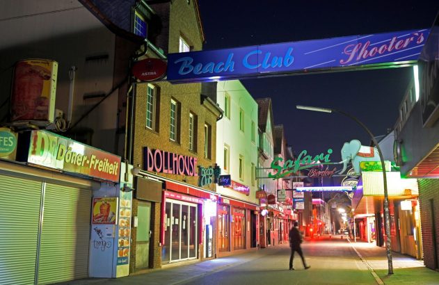 Rua noturna com lojas vibrantes, letreiros neon de 'Beach Club', 'DOLLHOUSE' e 'Döner-Freiheit'. Pessoa caminha.