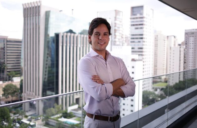 Homem sorrindo, braços cruzados, em sacada com prédios modernos ao fundo.