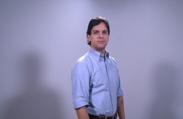 Homem de camisa azul, mangas arregaçadas, tatuagem no braço, olhando para câmera, em fundo cinza.