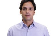 Homem moreno usando camisa azul-claro com logo polo, de frente, em fundo branco liso.