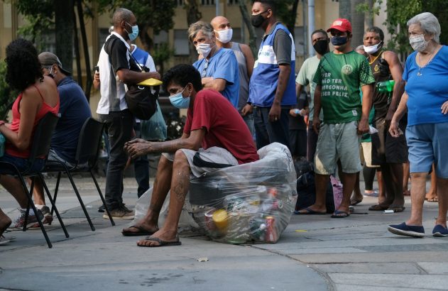 Grupo de pessoas diversas, com máscaras, em fila ao ar livre. Um homem senta sobre um saco transparente cheio de itens.
