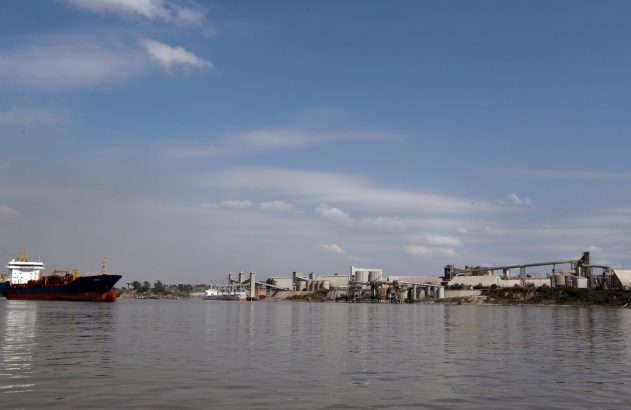 Navio no rio, com porto industrial e silos na margem direita sob céu azul.