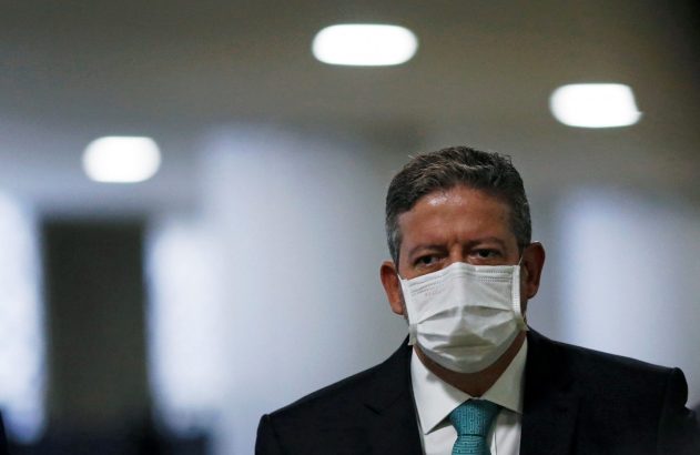 Homem de terno e gravata usando máscara branca, olhando para a câmera.