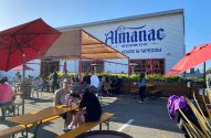 Cervejaria Almanac Beer Co. com área externa cheia de pessoas sentadas em mesas sob o sol.
