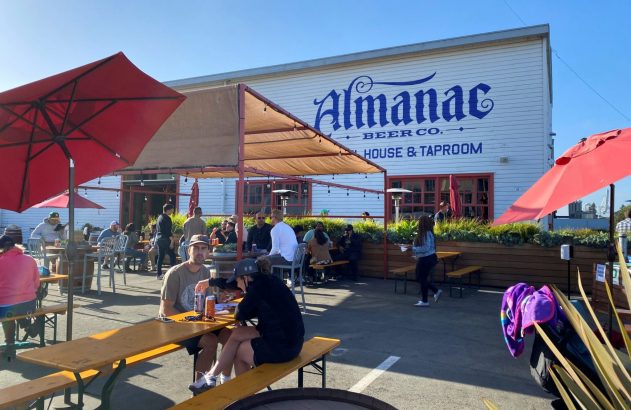 Cervejaria Almanac Beer Co. com área externa cheia de pessoas sentadas em mesas sob o sol.