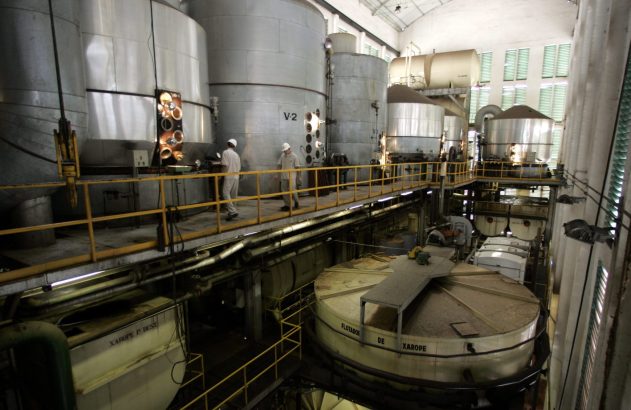 Fábrica de processamento de xarope com tanques industriais, maquinário e trabalhadores em plataformas.