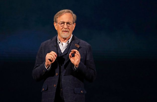 Steven Spielberg, de terno e óculos, gesticula ao falar contra fundo escuro.