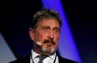 Retrato de John McAfee, homem com barba grisalha e microfone auricular, em fundo escuro com iluminação azul.