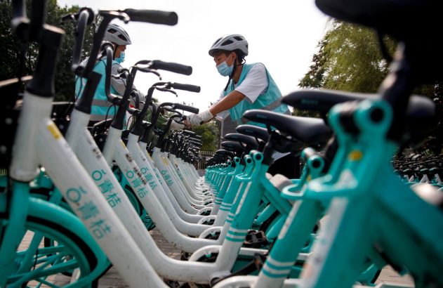 Trabalhadores com máscara e capacete organizam fileiras de bicicletas compartilhadas brancas e turquesas.