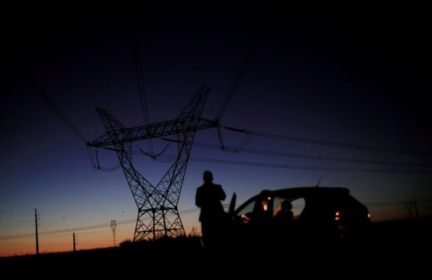 Silhueta de pessoa e carro ao anoitecer, com torre de energia e cabos contra o céu azul e laranja.