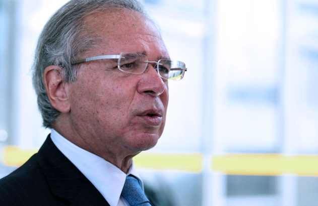 Homem de cabelo grisalho e óculos, em terno e gravata, boca semiaberta, falando em ambiente claro e desfocado.