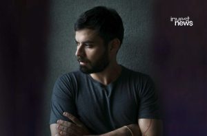 Homem barbudo de camisa cinza-escuro com braços cruzados olha para a esquerda, encostado em parede texturizada.