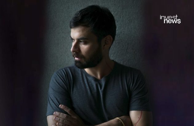 Homem barbudo de camisa cinza-escuro com braços cruzados olha para a esquerda, encostado em parede texturizada.