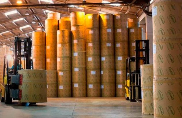 Empilhadeira em armazém transportando bobina; pilhas altas de bobinas de papelão ao fundo.