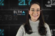 Mulher sorridente com óculos e suéter em estúdio de notícias financeiras com tela digital.