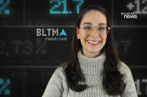Mulher sorridente com óculos e suéter em estúdio de notícias financeiras com tela digital.