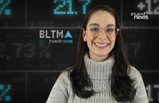 Mulher sorridente com óculos e suéter em estúdio de notícias financeiras com tela digital.
