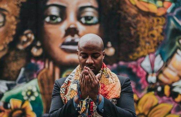 Homem de pele escura em oração, com terno e lenço estampado, contra um mural colorido com rosto feminino.