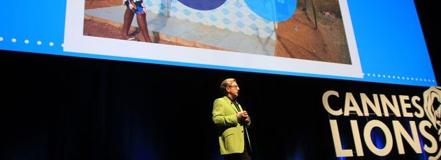 Palestrante em blazer verde no palco do Cannes Lions. Telão exibe imagem acima.