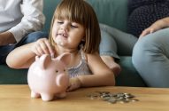 Menina deposita moedas em cofrinho rosa. Moedas espalhadas em mesa de madeira. Adultos no fundo.