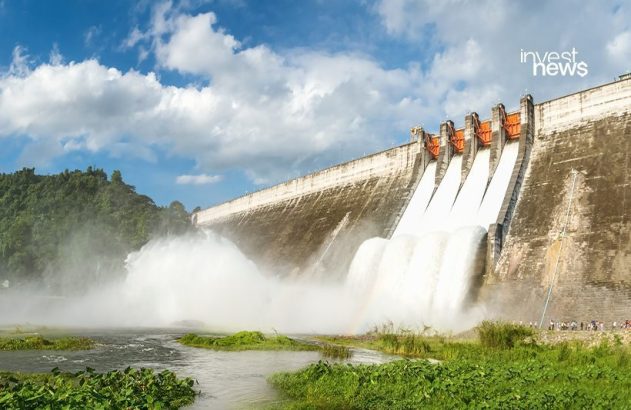 Barragem de concreto liberando muita água de várias comportas, criando névoa. Rio com vegetação e céu azul.