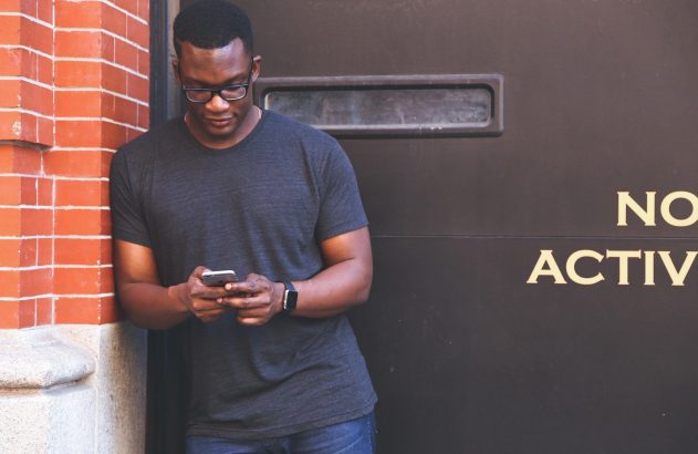 Homem de óculos olhando o smartphone, encostado em parede de tijolos e superfície escura.