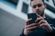 Homem segurando e usando um smartphone. As mãos e o aparelho estão em foco, o rosto do homem e o fundo estão desfocados.