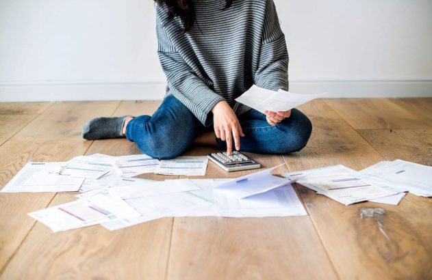 Pessoa sentada no chão de madeira, cercada por contas e papéis, usando uma calculadora para gerenciar finanças.