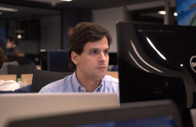 Homem de camisa azul olhando atentamente para um monitor de computador em um escritório.