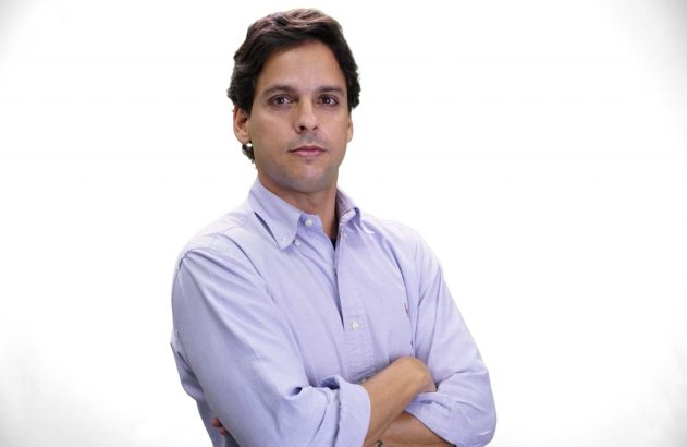 Homem de camisa azul, braços cruzados, olhando para a câmera em fundo branco.