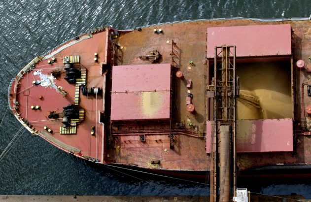 Vista aérea de navio graneleiro no porto, com porão aberto cheio de grãos e estrutura de carregamento.